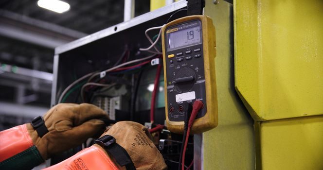 mains portant des gants de protection raccordant trois fils électriques marron, bleu et vert-jaune sur un bornier situé au dos d'une cuisinière électrique, tournevis posé à côté sur le sol de la cuisine
