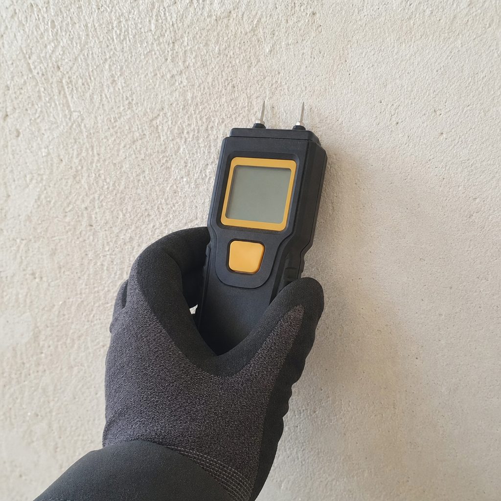 main gantée tenant un petit appareil testeur d'humidité avec deux pointes métalliques plantées dans un mur enduit, écran digital affichant un pourcentage