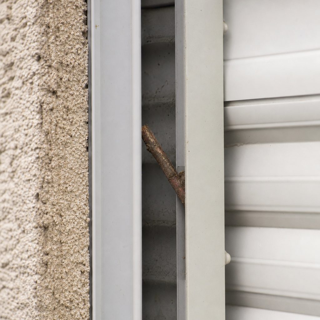 gros plan sur une glissière latérale de volet roulant avec une petite branche coincée à l'intérieur, lumière naturelle de jour